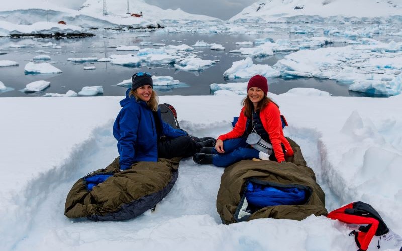 Zwei Frauen sitzen mit Schlafsäcken im Schnee auf der antarktischen Halbinsel während ihrer Kreuzfahrt.
